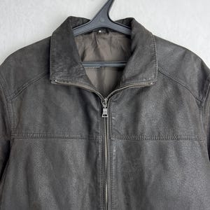 Puede incluir: Chaqueta de cuero marr&oacute;n con cremallera y cuello. La chaqueta est&aacute; colgada de una percha negra sobre un fondo blanco liso. La chaqueta tiene un aspecto texturizado.