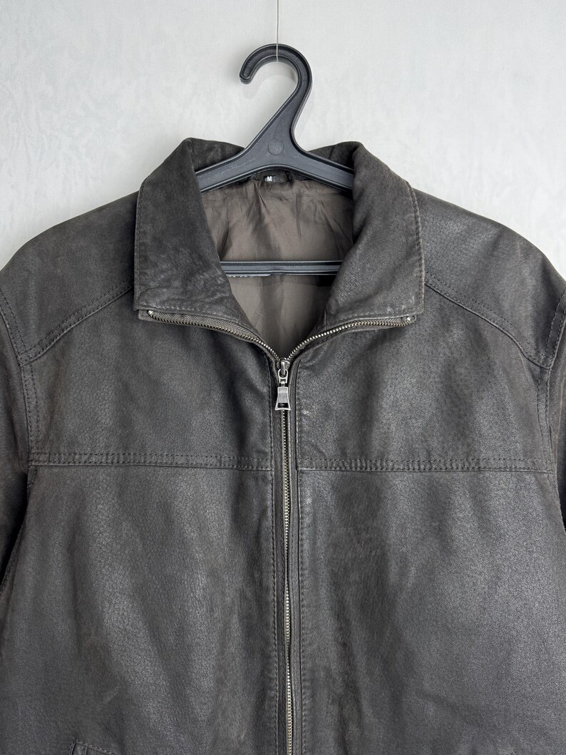 Puede incluir: Chaqueta de cuero marr&oacute;n con cremallera y cuello. La chaqueta est&aacute; colgada de una percha negra sobre un fondo blanco liso. La chaqueta tiene un aspecto texturizado.