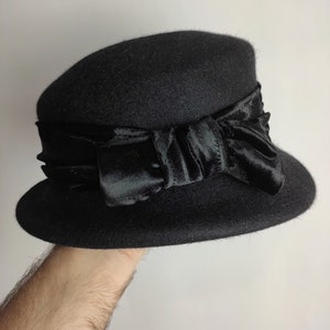 May include: A black cloche hat with a wide brim and a satin ribbon bow. The hat is made of a felt-like material and has a classic, vintage design. The bow is a prominent feature, adding a touch of elegance to the hat.
