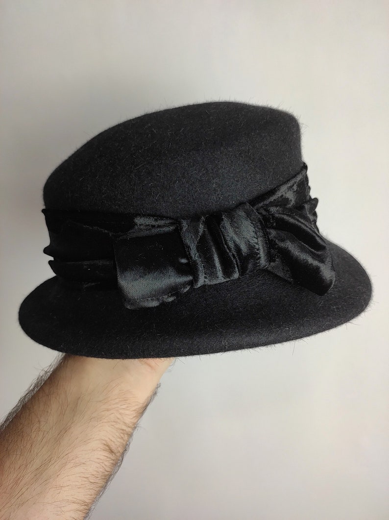 May include: A black cloche hat with a wide brim and a satin ribbon bow. The hat is made of a felt-like material and has a classic, vintage design. The bow is a prominent feature, adding a touch of elegance to the hat.