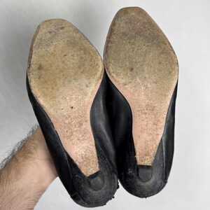 Puede incluir: Un par de zapatos de cuero negro con suelas desgastadas y tacones peque&ntilde;os. Los zapatos se sostienen sobre un fondo blanco, mostrando la parte inferior del calzado. Los tacones miden aproximadamente 3,8 cm de alto.