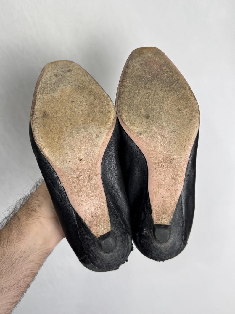 Puede incluir: Un par de zapatos de cuero negro con suelas desgastadas y tacones peque&ntilde;os. Los zapatos se sostienen sobre un fondo blanco, mostrando la parte inferior del calzado. Los tacones miden aproximadamente 3,8 cm de alto.