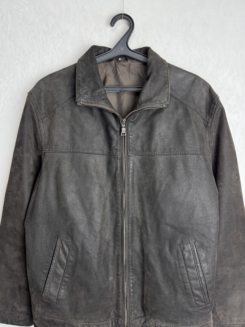 Puede incluir: Chaqueta de cuero marr&oacute;n oscuro con cierre de cremallera y dos bolsillos delanteros. La chaqueta est&aacute; colgada de una percha negra. El cuero tiene un aspecto texturado.