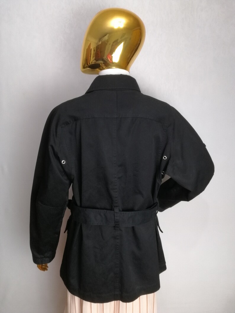 May include: Black belted jacket with long sleeves and a collar. The jacket has two pockets on the front and two on the sides.