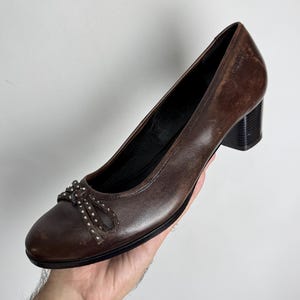 Puede incluir: Zapatos de tac&oacute;n de cuero marr&oacute;n con punta redondeada y un peque&ntilde;o detalle de lazo adornado con tachuelas plateadas. Los tacones son de color marr&oacute;n oscuro, estilo bloque. Los zapatos son sostenidos por una mano.