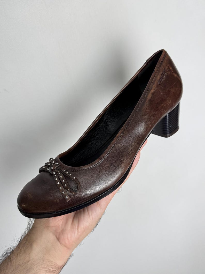 Puede incluir: Zapatos de tac&oacute;n de cuero marr&oacute;n con punta redondeada y un peque&ntilde;o detalle de lazo adornado con tachuelas plateadas. Los tacones son de color marr&oacute;n oscuro, estilo bloque. Los zapatos son sostenidos por una mano.