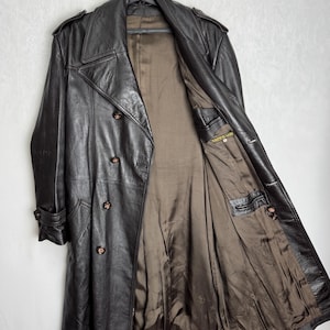 May include: A dark brown leather trench coat with a double-breasted design, hanging on a black hanger. The coat features a notched collar, button closures, and a brown interior lining. The coat has two front pockets and buttoned cuffs.