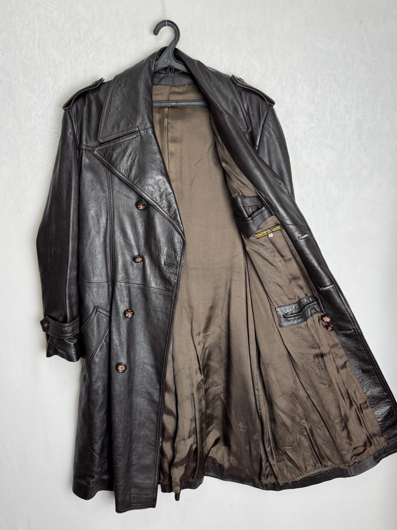 May include: A dark brown leather trench coat with a double-breasted design, hanging on a black hanger. The coat features a notched collar, button closures, and a brown interior lining. The coat has two front pockets and buttoned cuffs.