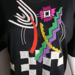 May include: Black t-shirt with a colourful geometric design embroidered on the front. The design features white, red, yellow, green, purple and pink thread.