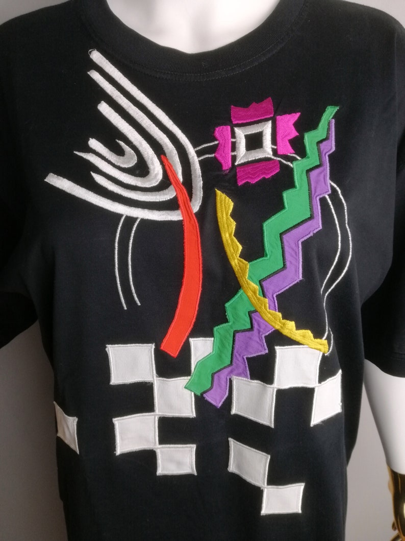 May include: Black t-shirt with a colourful geometric design embroidered on the front. The design features white, red, yellow, green, purple and pink thread.