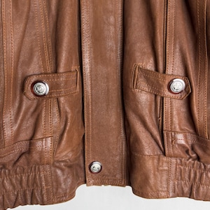 May include: Close-up of a brown leather jacket. The jacket features two front pockets with silver button closures. Vertical stitching details run down the front panels. The jacket has a ribbed hem and cuffs, and a button closure at the center.