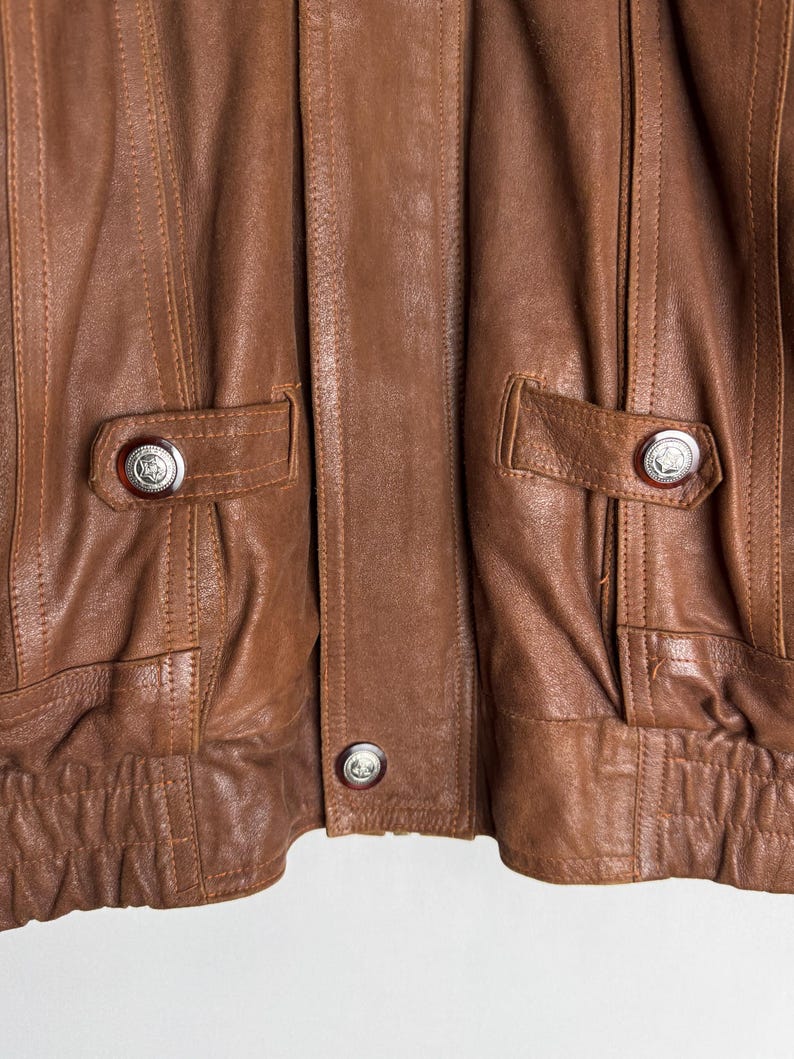 May include: Close-up of a brown leather jacket. The jacket features two front pockets with silver button closures. Vertical stitching details run down the front panels. The jacket has a ribbed hem and cuffs, and a button closure at the center.