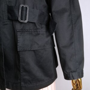 May include: Black utility jacket with a belt and a large pocket on the front. The jacket has a relaxed fit and is made of a durable fabric.