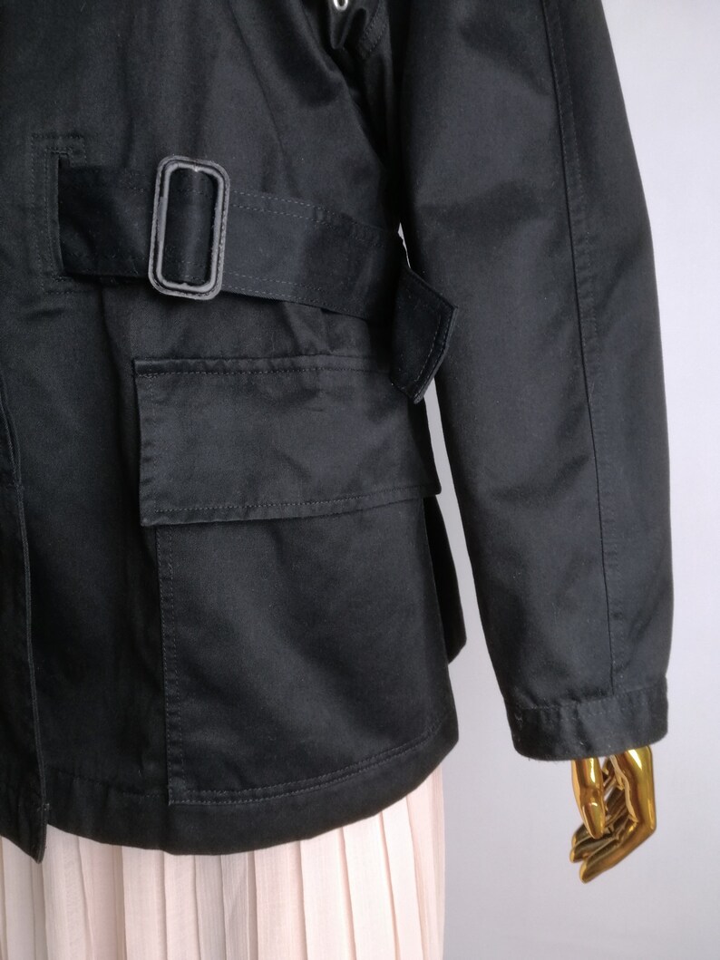 May include: Black utility jacket with a belt and a large pocket on the front. The jacket has a relaxed fit and is made of a durable fabric.