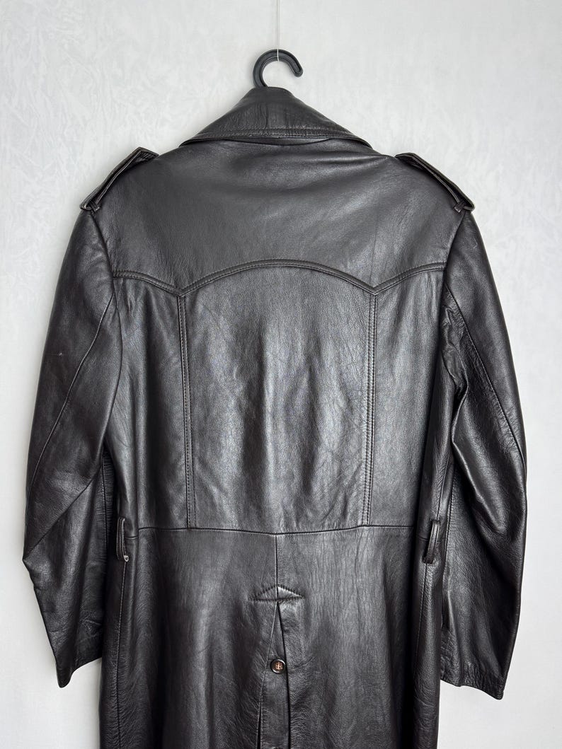 May include: Black leather coat hanging on a black hanger. The coat has a classic design with a notched collar, shoulder epaulets, and a button closure. The coat is made of a smooth, shiny leather material.