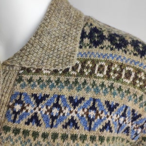 May include: Close-up of a beige wool sweater with a patterned design. The sweater features a textured collar and a detailed pattern in shades of blue, green, and brown. The overall style is a classic, warm knit.