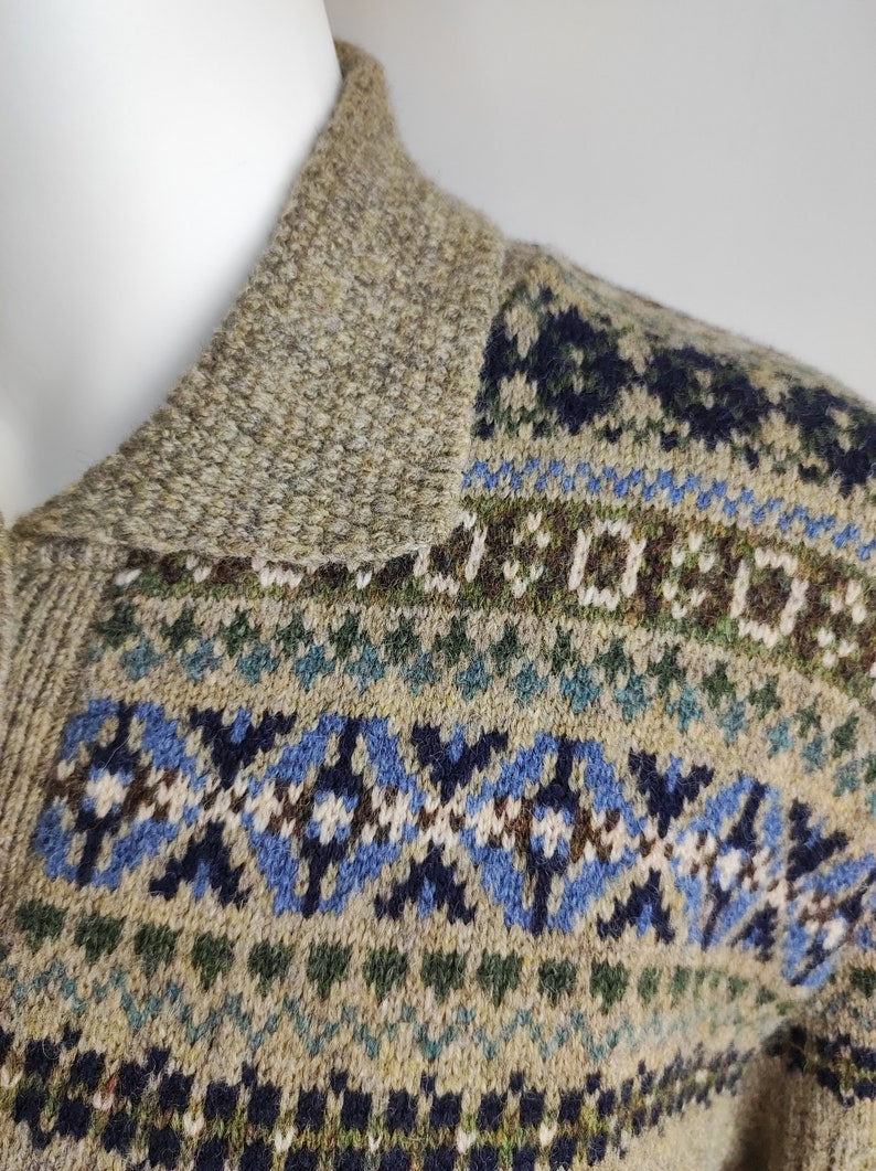 May include: Close-up of a beige wool sweater with a patterned design. The sweater features a textured collar and a detailed pattern in shades of blue, green, and brown. The overall style is a classic, warm knit.