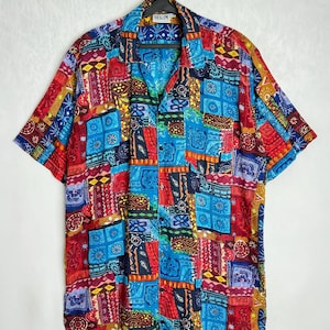 May include: A short-sleeved button-down shirt with a vibrant patchwork design. The shirt features a mix of red, blue, yellow, and purple patterns, including geometric shapes and floral motifs. The label reads "BEGUM".