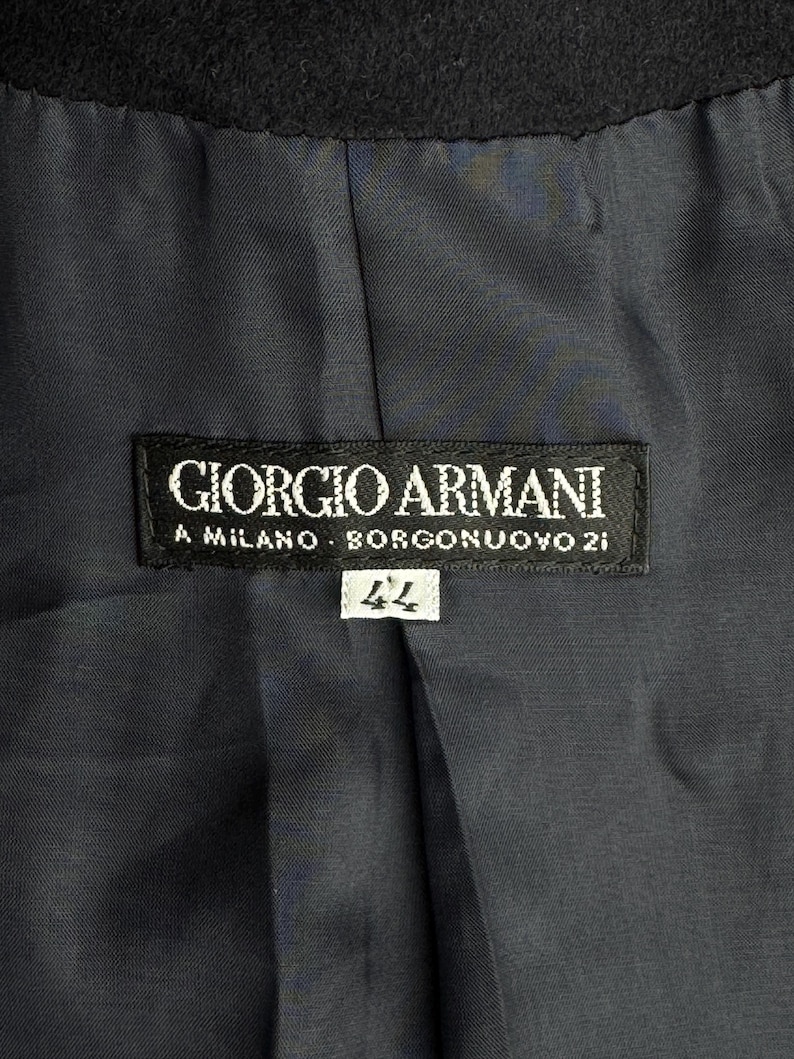 Pode incluir: Uma etiqueta preta Giorgio Armani com texto branco que diz "Giorgio Armani, A Milano, Borgonuvo 21" e uma etiqueta branca com o n&uacute;mero "44" nela.