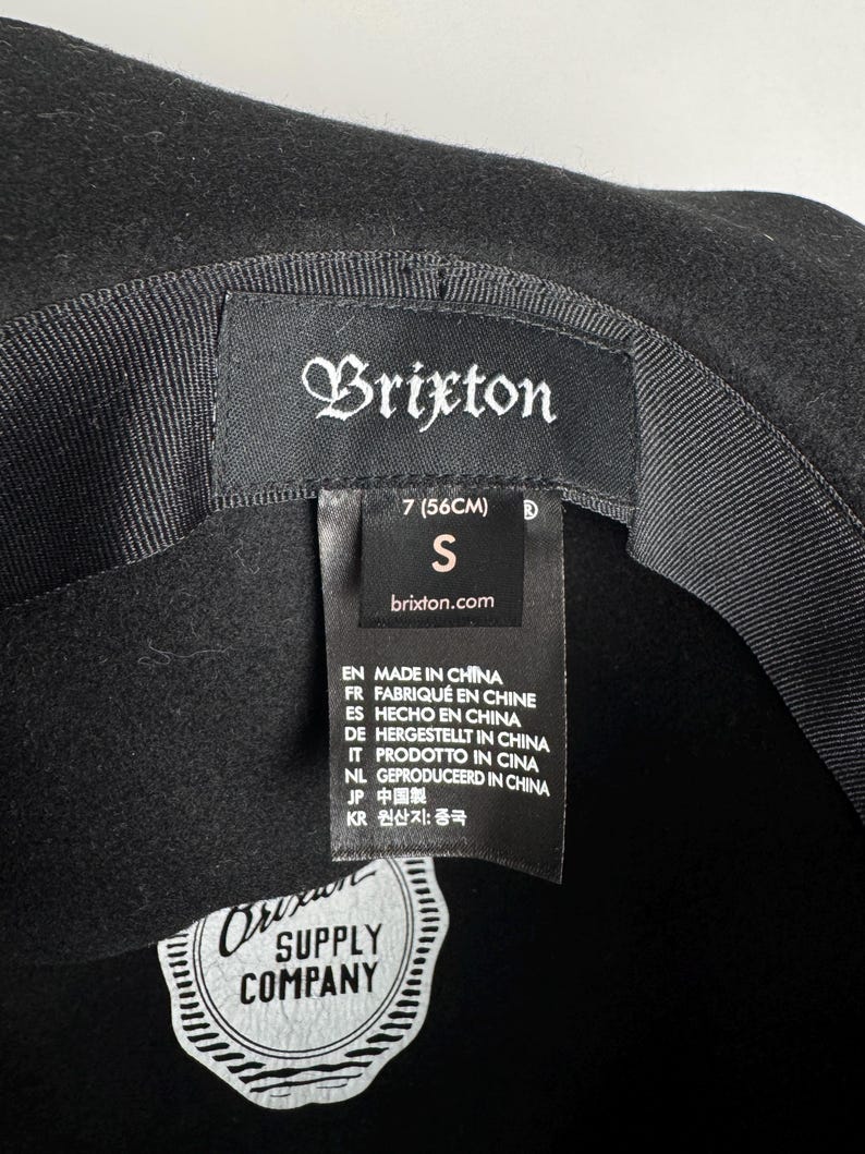 Puede incluir: Sombrero fedora de fieltro negro con una cinta negra y una etiqueta de Brixton Supply Company. El sombrero es talla 7 (56 cm) y talla S.