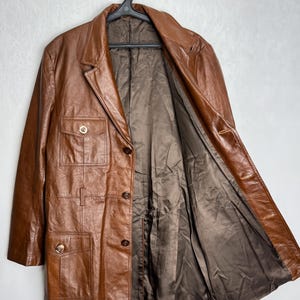 May include: A brown leather jacket with a classic design. The jacket features a notched collar, button closures, and multiple pockets. The interior lining is a darker brown color. The jacket is hanging on a black hanger.