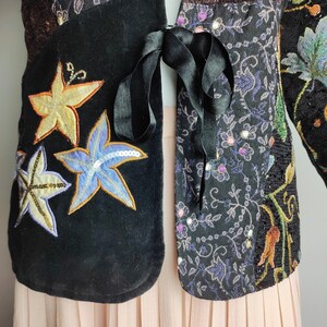 May include: A black velvet jacket with a patchwork design featuring embroidered flowers and sequins. The jacket has a tie closure and is worn over a pink pleated skirt.