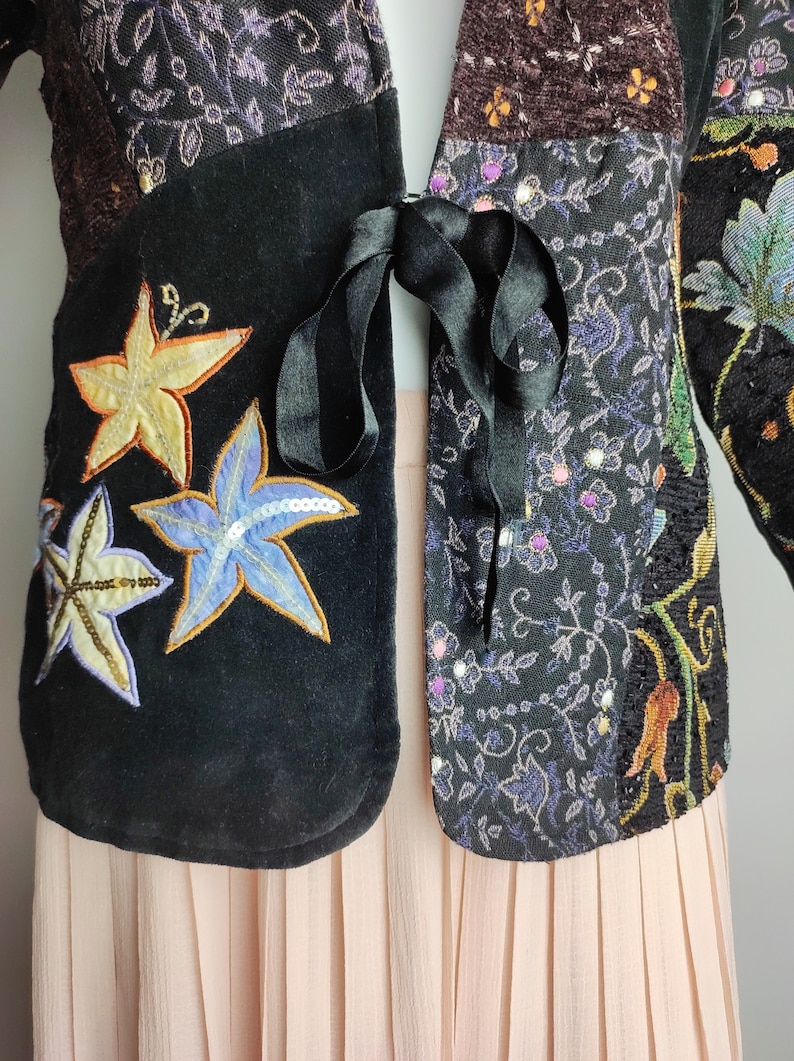 May include: A black velvet jacket with a patchwork design featuring embroidered flowers and sequins. The jacket has a tie closure and is worn over a pink pleated skirt.