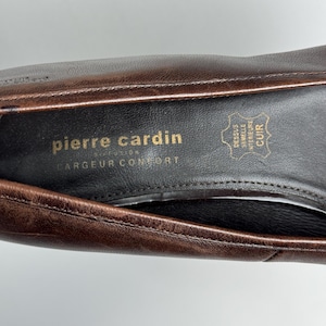 Puede incluir: Primer plano del interior de un zapato de cuero marr&oacute;n. La plantilla presenta el logotipo "pierre cardin" en letras doradas, junto con texto en franc&eacute;s. El interior del zapato es negro y el cuero tiene un aspecto texturizado.