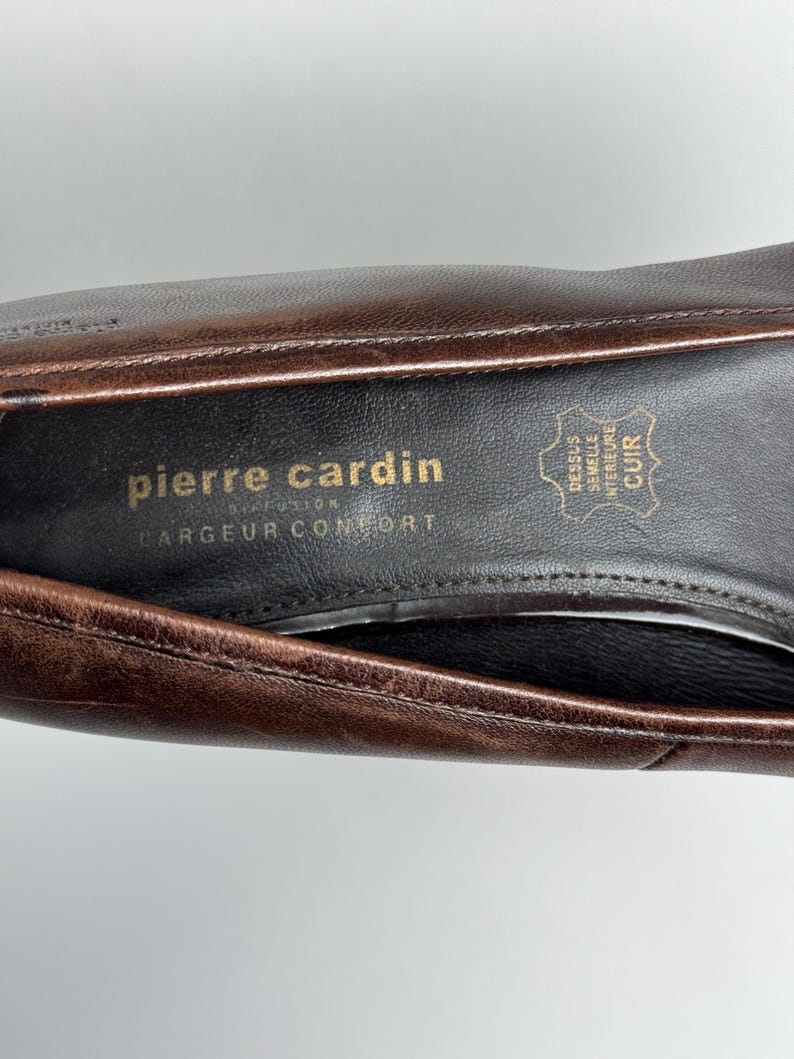 Puede incluir: Primer plano del interior de un zapato de cuero marr&oacute;n. La plantilla presenta el logotipo "pierre cardin" en letras doradas, junto con texto en franc&eacute;s. El interior del zapato es negro y el cuero tiene un aspecto texturizado.