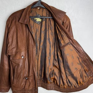 May include: A brown leather jacket with a zipper closure, two front pockets, and a collared neckline. The jacket has a brown paisley patterned lining and the brand name "Laval Conti" is visible on the inside.