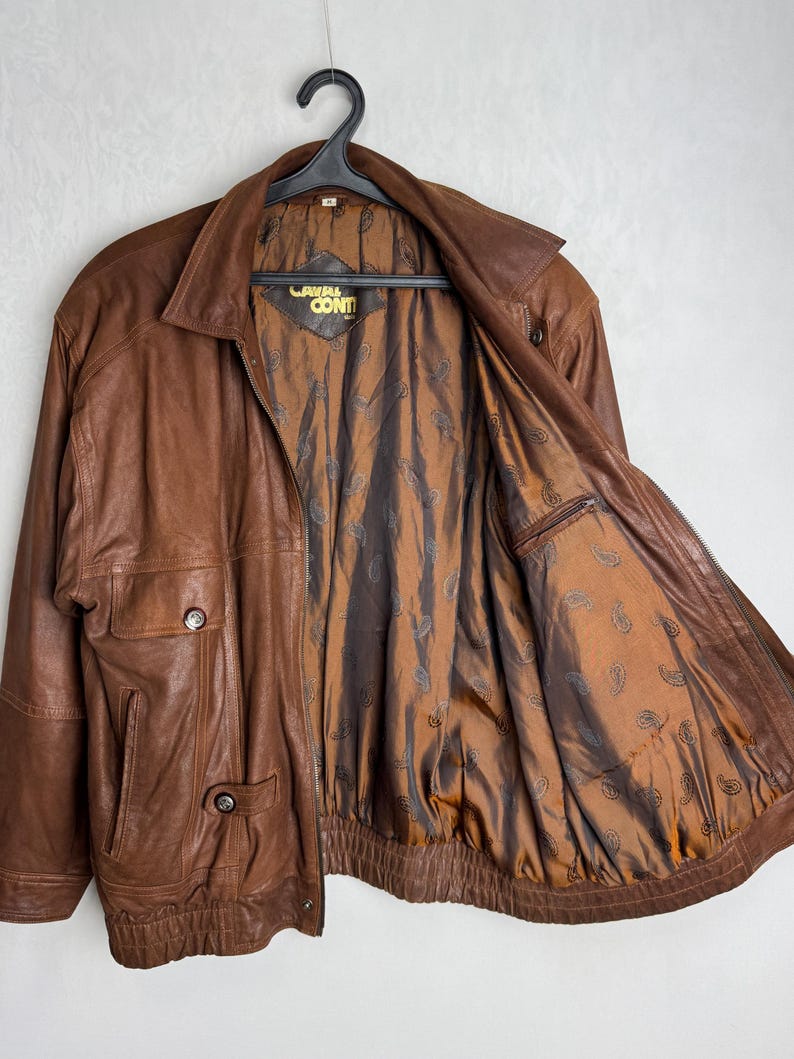 May include: A brown leather jacket with a zipper closure, two front pockets, and a collared neckline. The jacket has a brown paisley patterned lining and the brand name "Laval Conti" is visible on the inside.