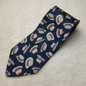 May include: A navy blue silk tie with a repeating pattern of white and purple hats. The hats have a variety of designs, including stripes, polka dots, and solid colors.