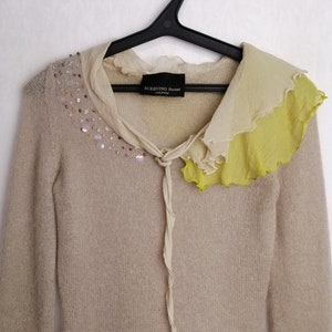 Puede incluir: Un su&eacute;ter de punto beige con un cuello de volantes en tonos beige y verde. El cuello tiene un detalle de corbata y el su&eacute;ter est&aacute; adornado con lentejuelas iridiscentes. El su&eacute;ter tiene la etiqueta "SCERVINO Street" en la parte delantera.