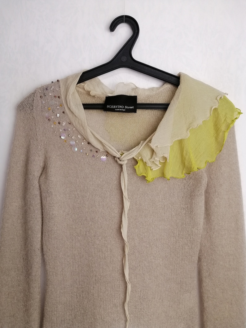 Puede incluir: Un su&eacute;ter de punto beige con un cuello de volantes en tonos beige y verde. El cuello tiene un detalle de corbata y el su&eacute;ter est&aacute; adornado con lentejuelas iridiscentes. El su&eacute;ter tiene la etiqueta "SCERVINO Street" en la parte delantera.