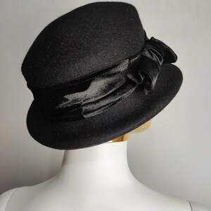 May include: A black cloche hat with a velvet ribbon and bow. The hat has a rounded crown and a slightly turned-down brim. The velvet ribbon is wrapped around the base of the crown and tied into a bow on the side. The hat is displayed on a mannequin head.