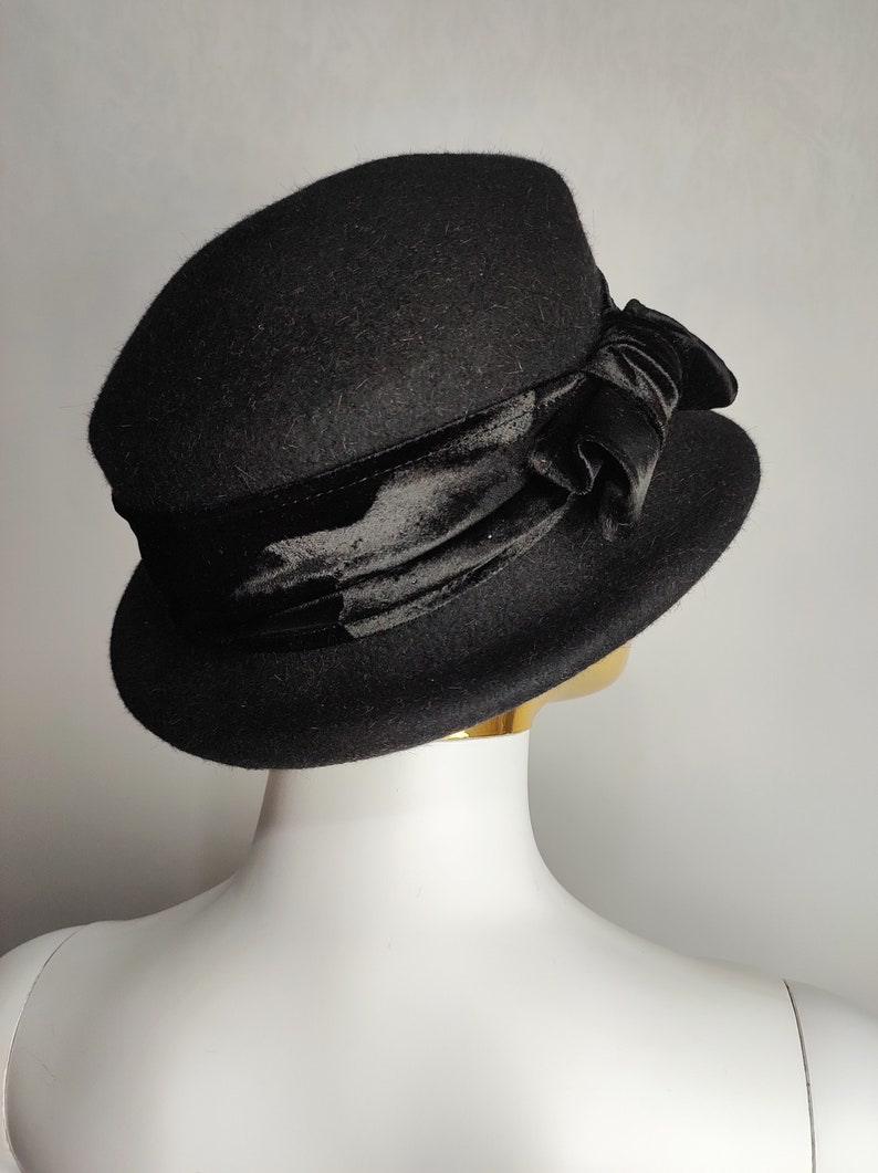 May include: A black cloche hat with a velvet ribbon and bow. The hat has a rounded crown and a slightly turned-down brim. The velvet ribbon is wrapped around the base of the crown and tied into a bow on the side. The hat is displayed on a mannequin head.