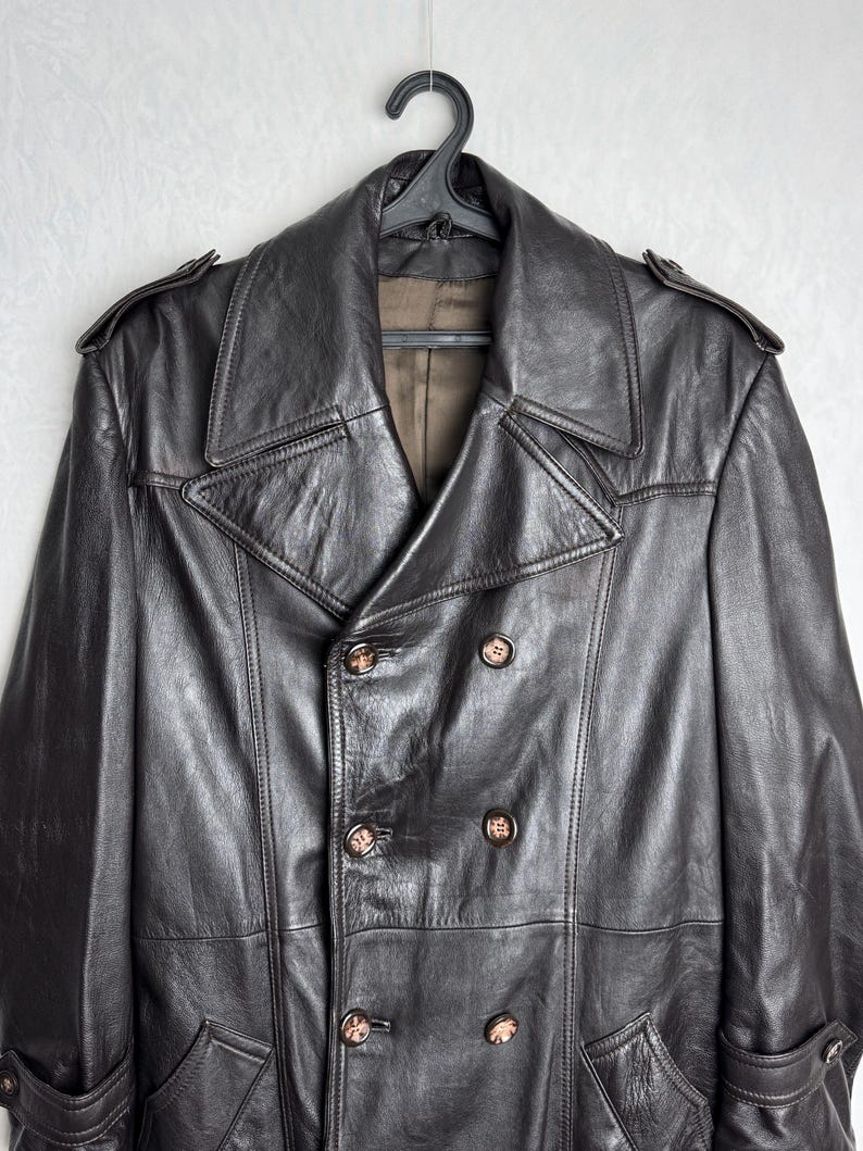 May include: A dark brown leather coat with a double-breasted design, featuring large lapels and decorative buttons. The coat is hanging on a black hanger, showcasing its classic style and texture.
