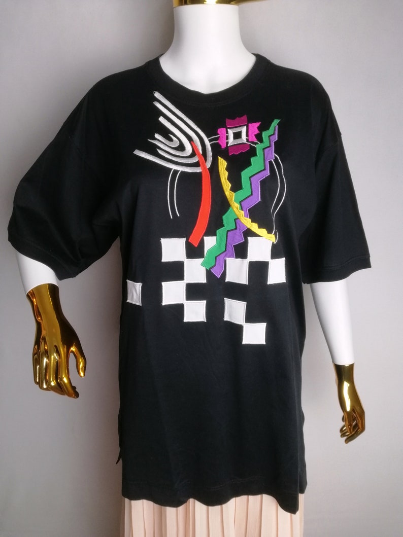May include: Black t-shirt with a colourful abstract design featuring geometric shapes, lines, and a chequered pattern.
