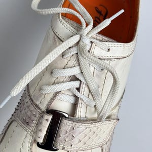 K&ouml;nnte beinhalten: Nahaufnahme eines wei&szlig;en Ledersneakers mit wei&szlig;en Schn&uuml;rsenkeln. Der Schuh hat einen Riemen mit Schlangenmuster und einer silbernen Schnalle. Die Innenseite ist orange. Vertikale N&auml;hte zieren das Design des Schuhs.