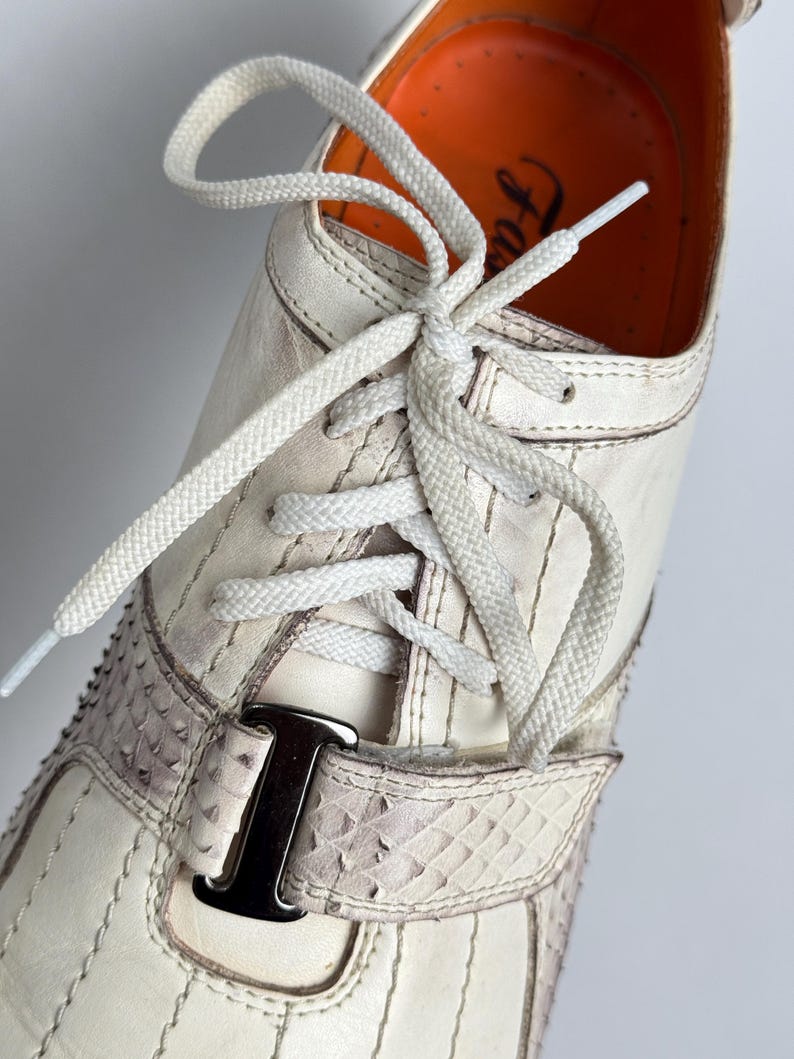 K&ouml;nnte beinhalten: Nahaufnahme eines wei&szlig;en Ledersneakers mit wei&szlig;en Schn&uuml;rsenkeln. Der Schuh hat einen Riemen mit Schlangenmuster und einer silbernen Schnalle. Die Innenseite ist orange. Vertikale N&auml;hte zieren das Design des Schuhs.