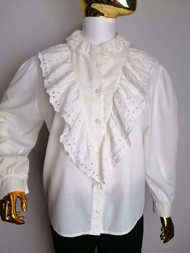 Puede incluir: Blusa blanca de manga larga con un gran cuello con volantes y detalles de ojal. La blusa tiene un cierre de botones en la parte delantera.