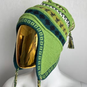 Puede incluir: Un gorro de punto verde lima con orejeras, que presenta patrones geométricos en verde oscuro, oliva y blanco. El gorro incluye lazos trenzados que cuelgan de las orejeras y una pequeña borla decorativa en la coronilla, sobre un maniquí dorado.