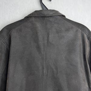 Puede incluir: Chaqueta de cuero marr&oacute;n oscuro, colgada de una percha negra. La chaqueta tiene un cuello cl&aacute;sico y una superficie texturizada. La parte trasera de la chaqueta muestra los detalles de las costuras.