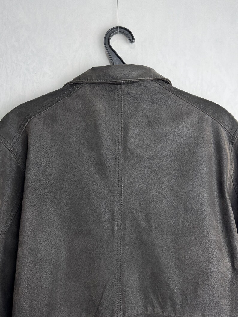 Puede incluir: Chaqueta de cuero marr&oacute;n oscuro, colgada de una percha negra. La chaqueta tiene un cuello cl&aacute;sico y una superficie texturizada. La parte trasera de la chaqueta muestra los detalles de las costuras.