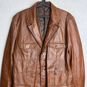 May include: A brown leather jacket with a classic design, featuring a notched collar, four front pockets, and gold-colored buttons. The jacket is hanging on a black hanger against a plain white background. The jacket appears to be a medium size.