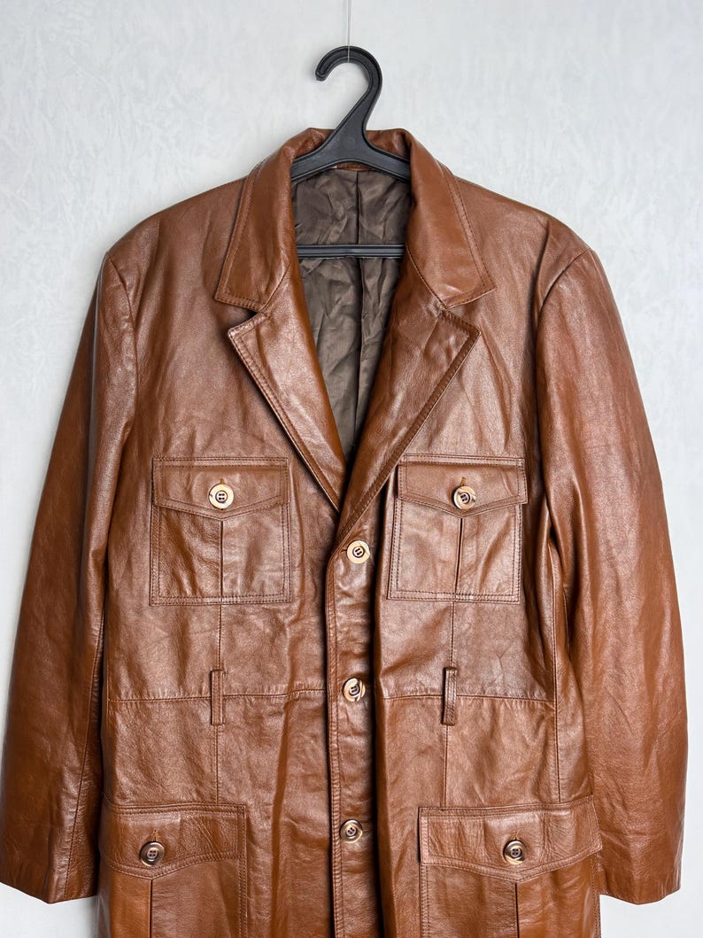 May include: A brown leather jacket with a classic design, featuring a notched collar, four front pockets, and gold-colored buttons. The jacket is hanging on a black hanger against a plain white background. The jacket appears to be a medium size.