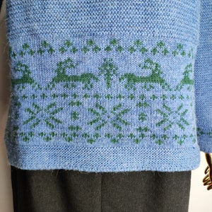 May include: A blue knitted jumper with a green and white pattern of trees and reindeer. The jumper has a ribbed hem and cuffs.