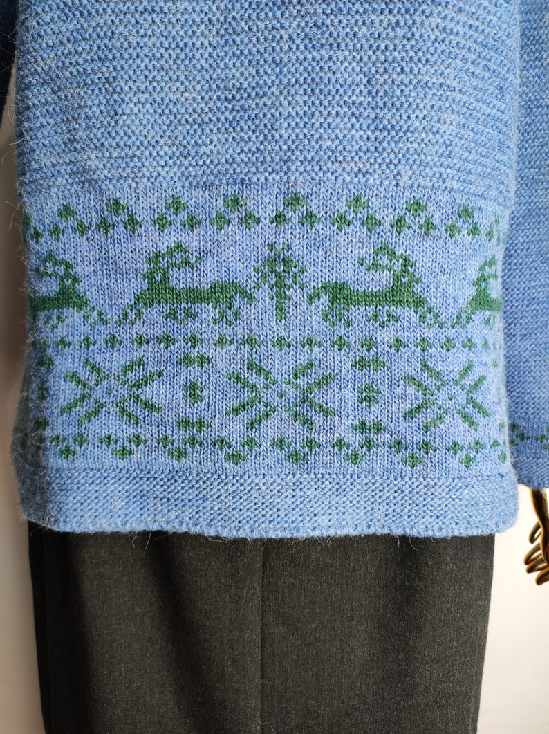 May include: A blue knitted jumper with a green and white pattern of trees and reindeer. The jumper has a ribbed hem and cuffs.