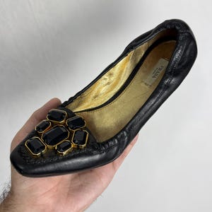 Puede incluir: Bailarina Prada de cuero negro con interior dorado. El zapato presenta un grupo de grandes piedras preciosas negras, rectangulares y octogonales, engastadas en un marco de metal dorado en la puntera. El logotipo de Prada es visible en el interior.