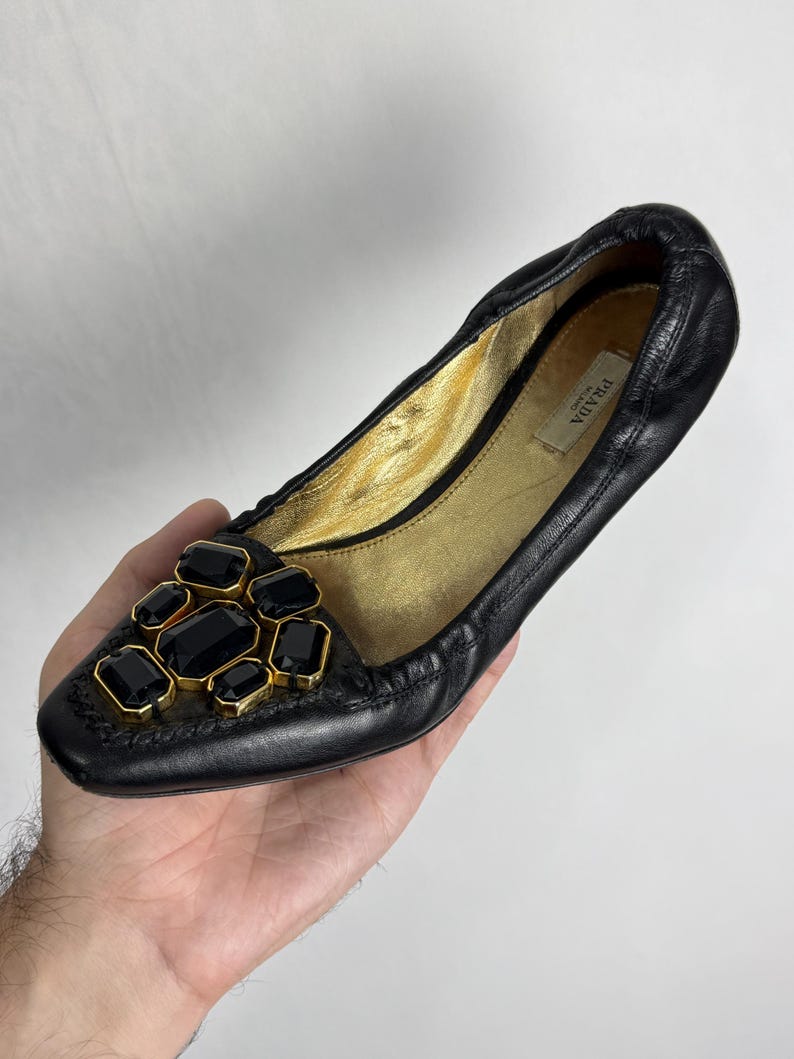 Puede incluir: Bailarina Prada de cuero negro con interior dorado. El zapato presenta un grupo de grandes piedras preciosas negras, rectangulares y octogonales, engastadas en un marco de metal dorado en la puntera. El logotipo de Prada es visible en el interior.